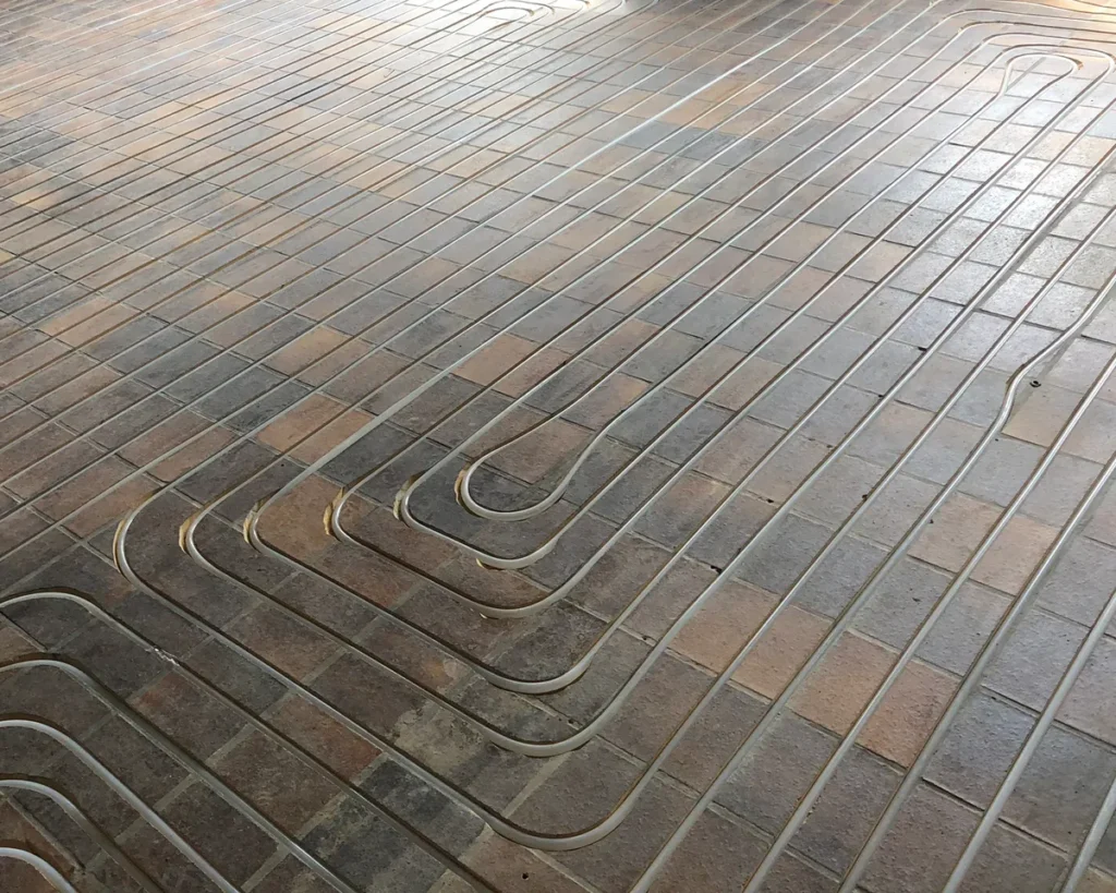 Op de foto liggen flexibele kunststofbuizen strak in de tegelvloer verwerkt voor vloerverwarming. Dit betreft een watergedragen vloerverwarmingsinstallatie onder bestaande of nieuwe tegels. Het hoofddoel is om via de hele vloer een gelijkmatige en energiezuinige verwarming te bieden. De buizen zijn nauwkeurig gebogen om in elke hoek te passen, de vaste tussenafstand garandeert een gelijkmatige warmteafgifte en het systeem werkt onzichtbaar onder de vloer zodat de leefruimte vrij blijft van radiatoren.